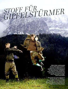 Zwei Männer in traditioneller bayerischer Kleidung vollführen einen Sprung vor einer malerischen Kulisse aus schneebedeckten Bergen, Wald und bewölktem Himmel. Der deutsche Text lautet "Stoff für Gipfelstürmer" am oberen Rand.