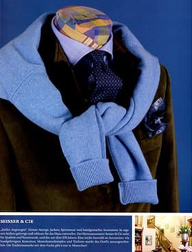 Eine Schaufensterpuppe trägt eine braune Jacke, ein blaues Hemd, eine marineblaue gepunktete Krawatte und einen hellblauen Pullover, der über die Schultern gezogen ist. Der Kopf der Schaufensterpuppe ist mit einer bunten, gemusterten Kappe bedeckt.