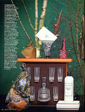 Ein Stillleben mit grünem Hintergrund, kahlen Ästen, einem Glasgeschirr mit sechs Gläsern und einer Karaffe, einer weißen Flasche, gefalteten Taschentüchern, einer braunen Tasche, Büchern und gemusterten Tüchern.