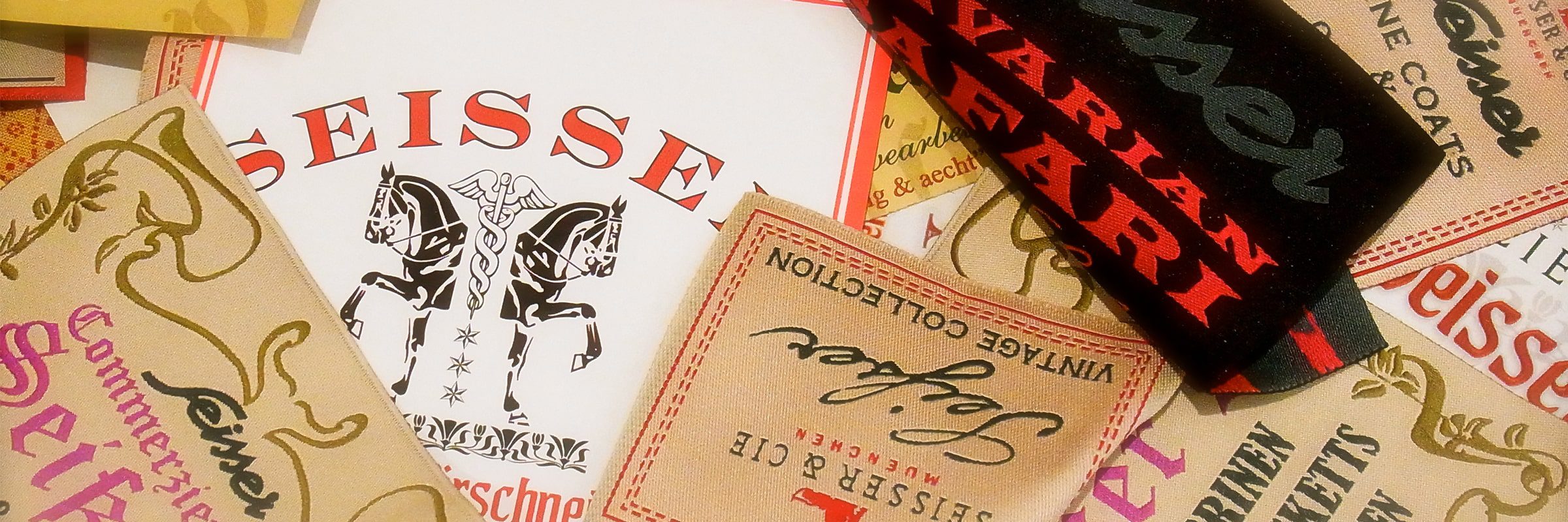 Eine Nahaufnahme verschiedener Kleidungsetiketten aus Stoff und Papier mit unterschiedlichen Schriftarten, Farben und Mustern, die sich teilweise überlappen. In der Mitte sind ein prominentes Pferdelogo und das Wort "SEISSE" zu sehen.