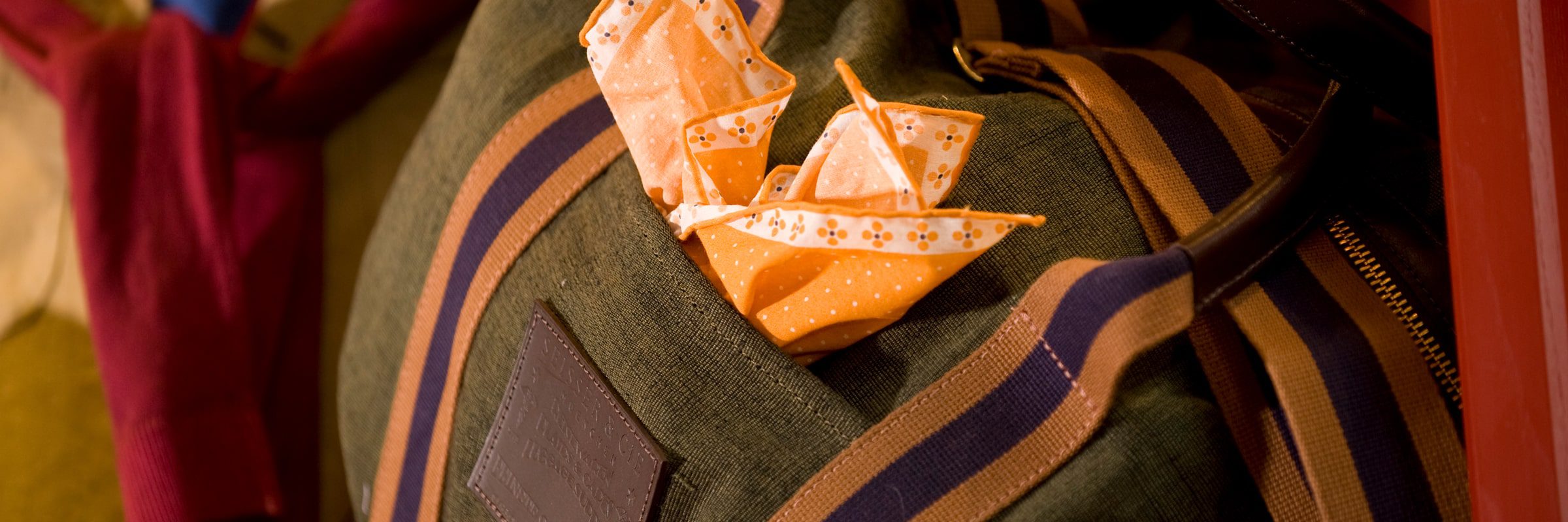Eine graue Tasche mit braun und blau gestreiften Trägern hat ein orange gemustertes Taschentuch in der Vordertasche. Im Hintergrund ist eine rote Krawatte zu sehen.