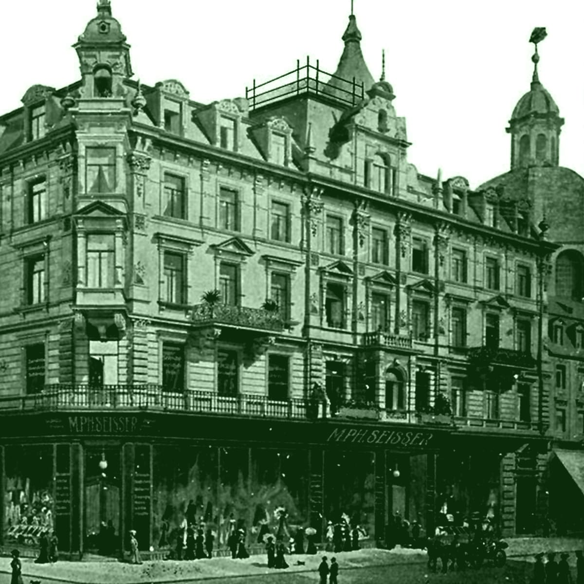 Ein historisches mehrstöckiges Gebäude mit verschnörkelten architektonischen Details und Geschäften auf Straßenebene, darunter "M. P. Belser", mit Menschen, die sich draußen versammelt haben; im Hintergrund ist ein weiteres Hochhaus zu sehen. Das Bild hat einen Grünstich.