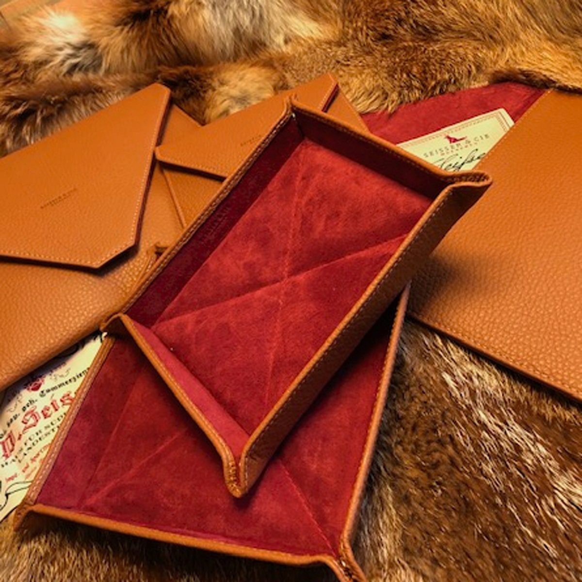 Mehrere braune Ledertabletts und Briefumschläge mit roter Wildlederinnenseite sind auf einer Pelzoberfläche ausgestellt, wobei in einigen von ihnen Papiere im Vintage-Stil teilweise sichtbar sind.