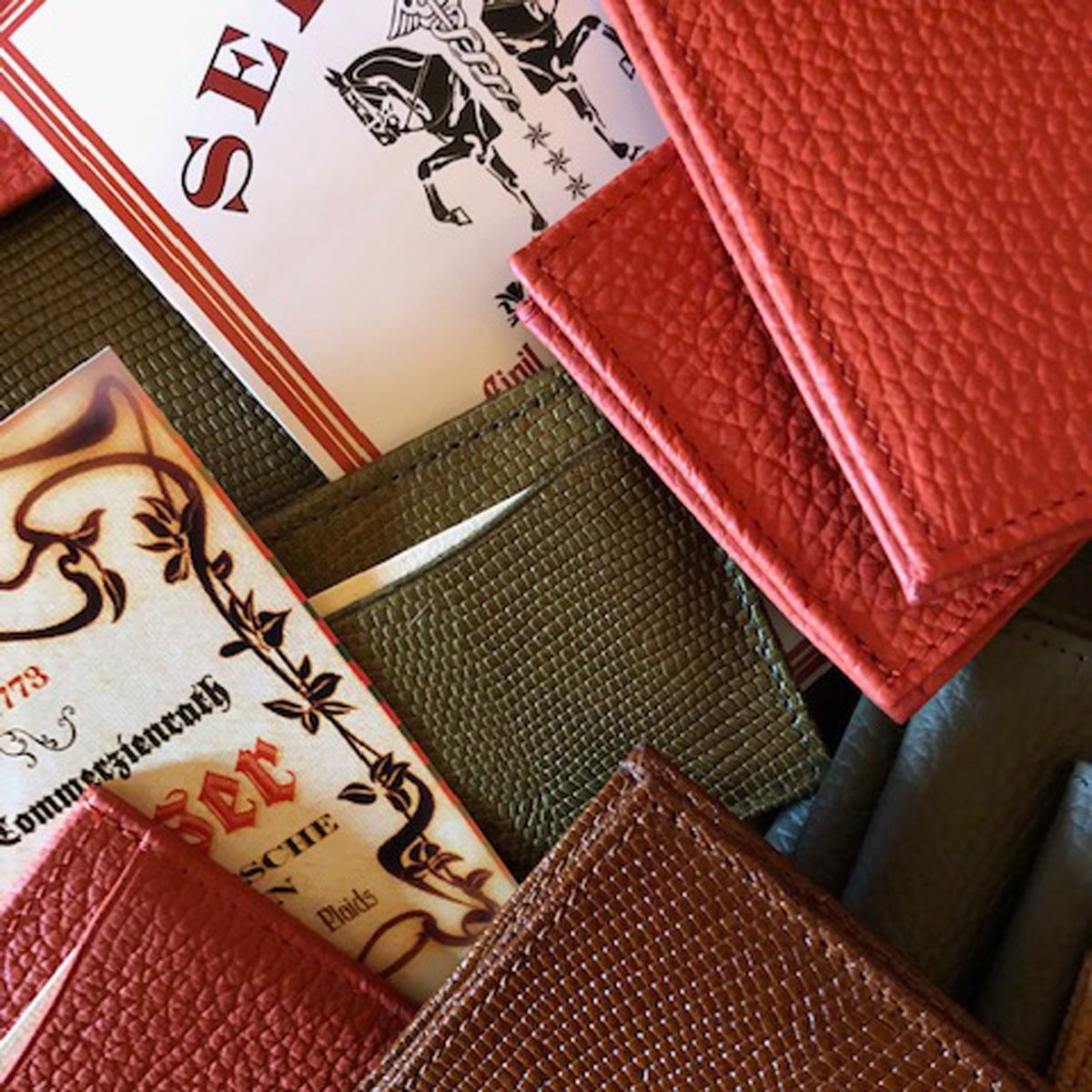 © Seisser & Cie. Muenchen GmbH Nahaufnahme von Brieftaschen aus strukturiertem Leder in Rot, Grün und Braun, die teilweise verzierte Papierkarten mit dekorativen Mustern und Text enthalten.