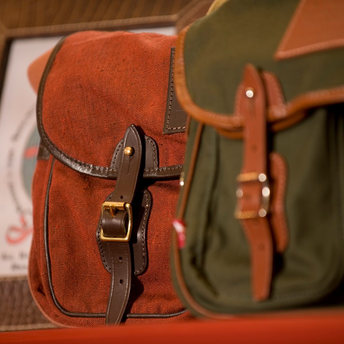 © Seisser & Cie. Muenchen GmbH Zwei Segeltuchtaschen mit Lederriemen und Schnallen, eine in Rotbraun, die andere in Grün, sind nebeneinander gestellt. Der Fokus liegt auf den Riemen und Schnallen. Der Hintergrund ist unscharf.