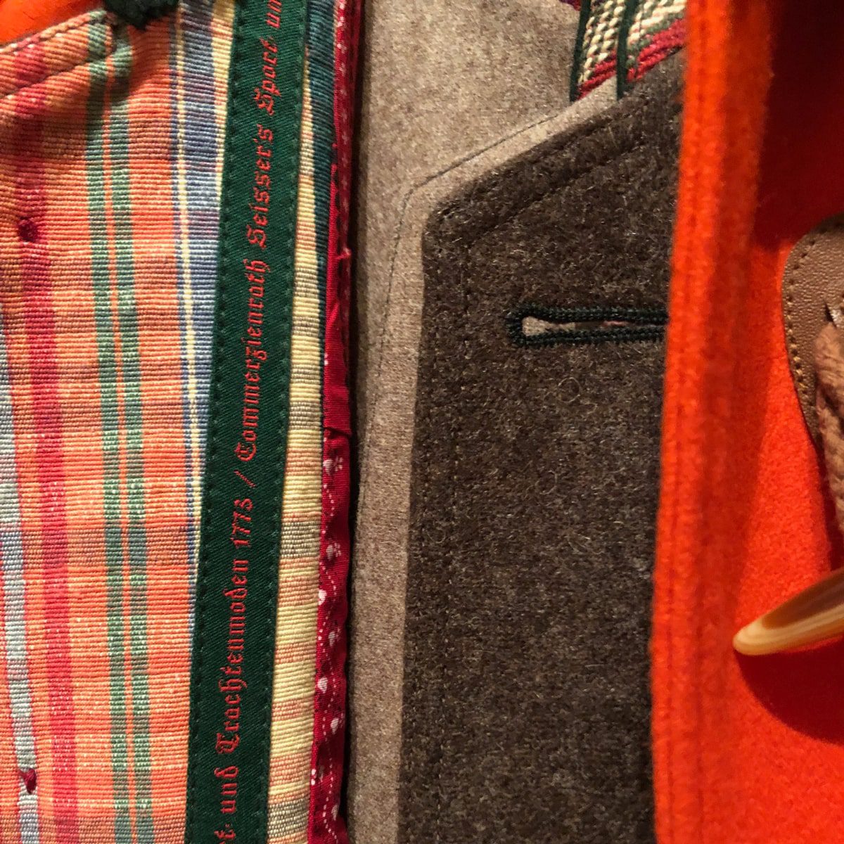 Nahaufnahme der Innenseite eines Mantels, die einen bunt karierten Stoff, graue Wolle und orangefarbenes Material zeigt. Am Rand des karierten Futters ist ein grüner Text in deutscher Sprache aufgestickt. Ein Holzbügel ist teilweise sichtbar.
