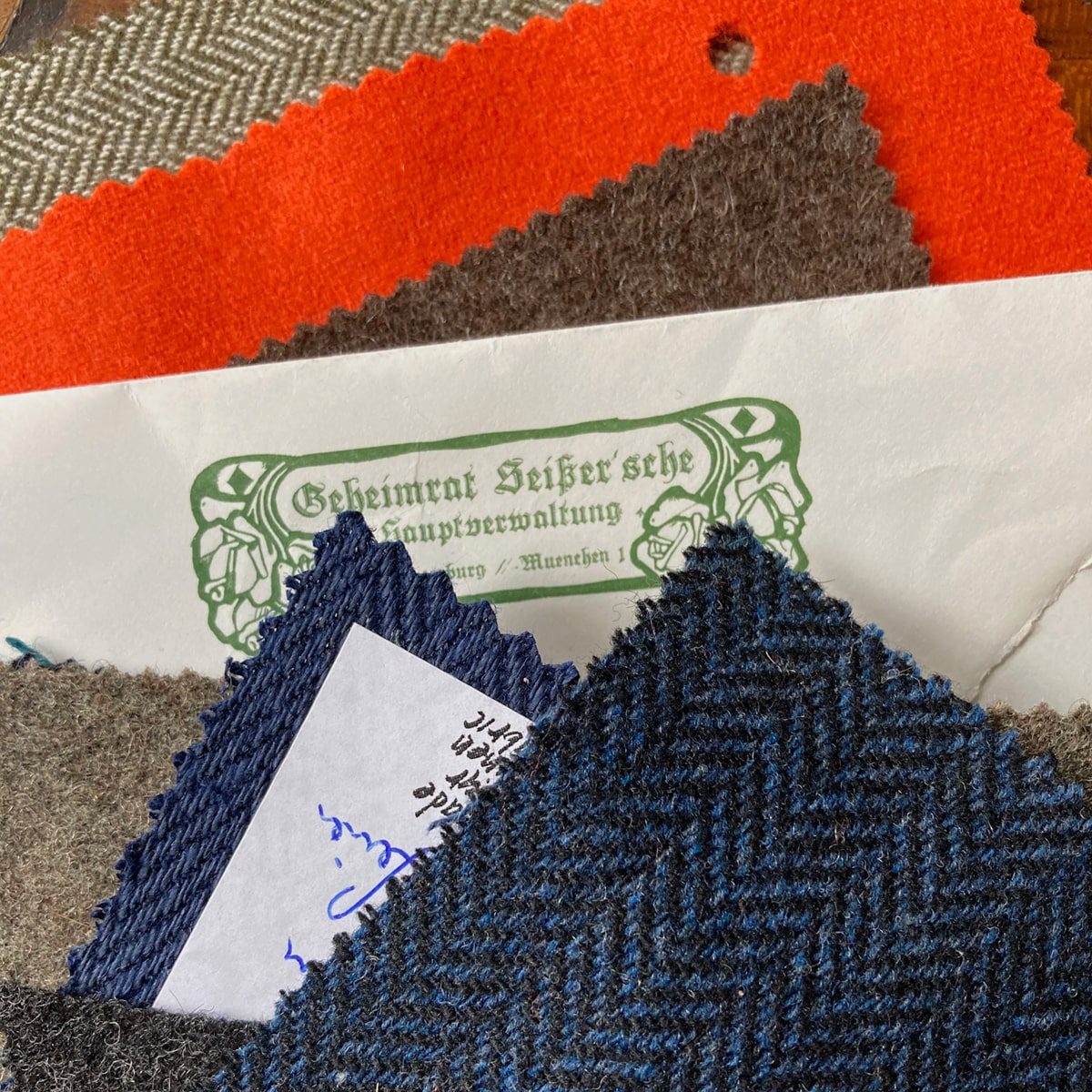 Mehrere Stoffmuster in verschiedenen Farben und Mustern, darunter Grau, Blau, Orange und Fischgrät, sind übereinandergelegt. Unter den Stoffen befindet sich ein teilweise sichtbarer Umschlag mit verschnörkeltem deutschen Text.