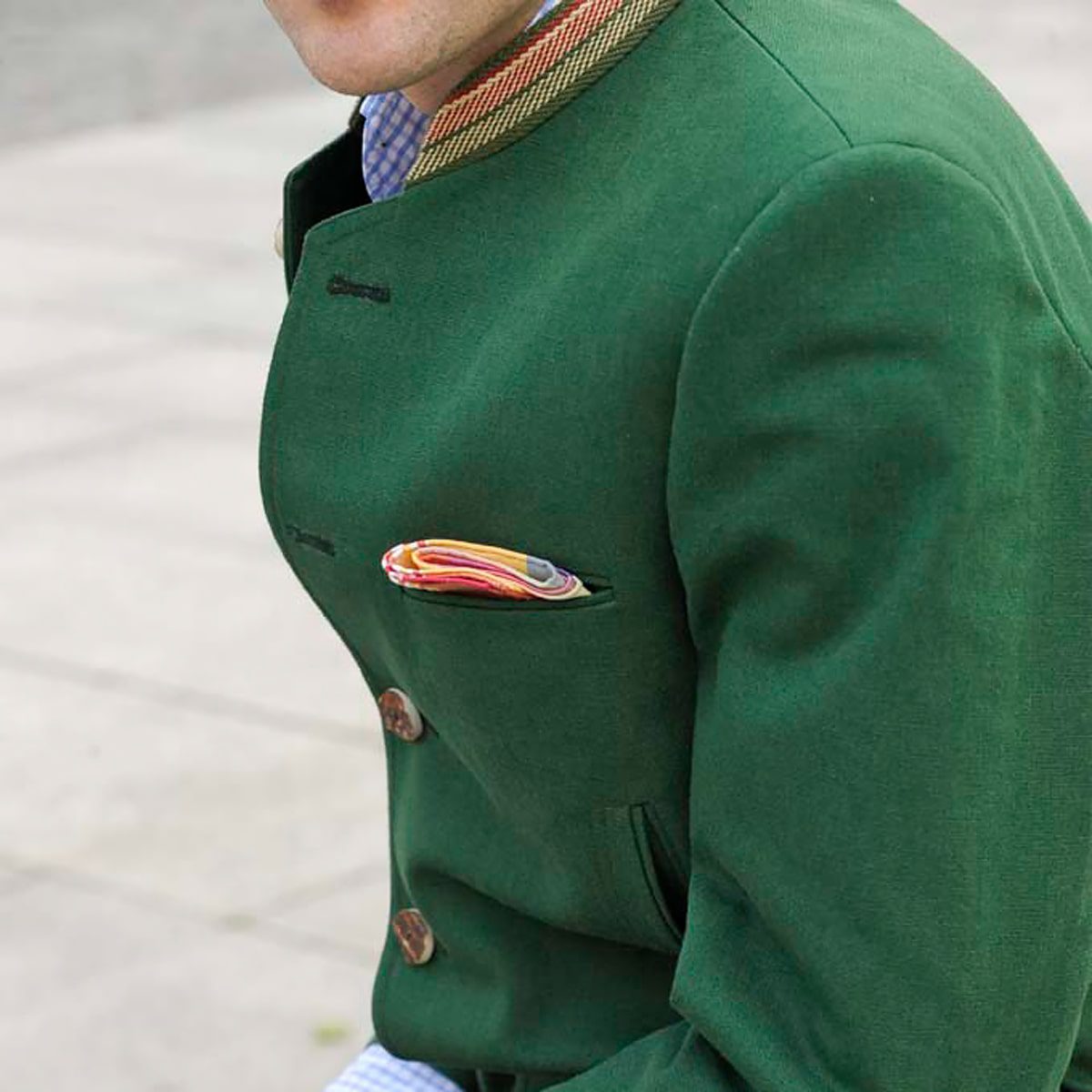 Eine Person, die eine grüne Jacke mit Zierknöpfen und ein buntes Einstecktuch trägt, wird von den Schultern abwärts, leicht zur Seite gedreht, im Freien auf einer gepflasterten Fläche gezeigt.