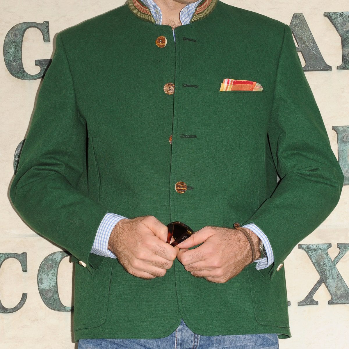 Eine Person, die eine grüne Jacke im traditionellen Stil mit Holzknöpfen und ein buntes Einstecktuch trägt, steht vor einer Wand mit teilweise sichtbaren Buchstaben. Nur der Oberkörper und die Hände sind sichtbar.