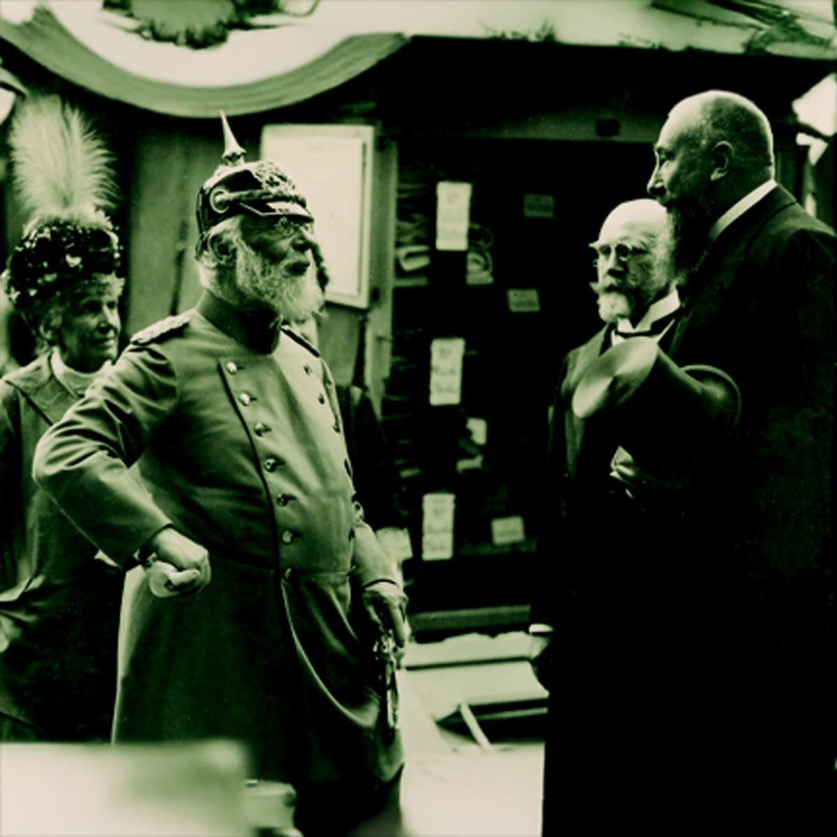 Vier Männer in historischen Gewändern stehen zusammen und unterhalten sich. Einer trägt eine verzierte Militäruniform mit Stachelhelm, die anderen formelle Anzüge; einer hat einen Federhut. Der Schauplatz scheint im Freien zu sein, möglicherweise bei einer Veranstaltung oder Versammlung.