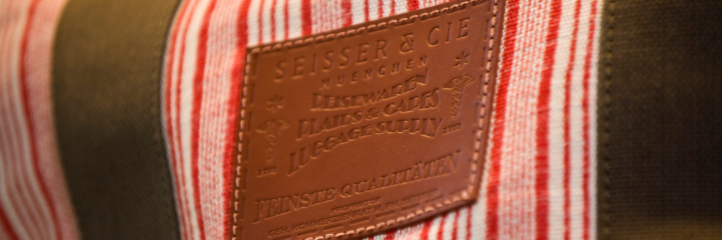 Eine Nahaufnahme eines Lederaufnähers mit geprägtem Text auf einer Stofftasche mit roten und weißen Längsstreifen und olivgrünen Akzenten. Auf dem Aufnäher steht: "Seisser & Cie München Handarbeit Plaids & Co. Luggage Supply".