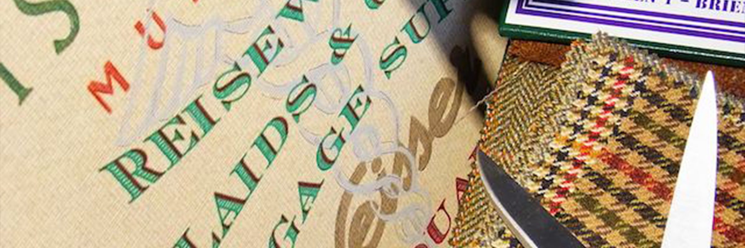 Nahaufnahme eines Stoffes mit den Wörtern "REISE", "LUGGAGE" und "SUIT" in Grün und Rot, daneben eine Schere, gemusterter Tweed-Stoff und ein Etikett, das in der oberen rechten Ecke teilweise sichtbar ist.