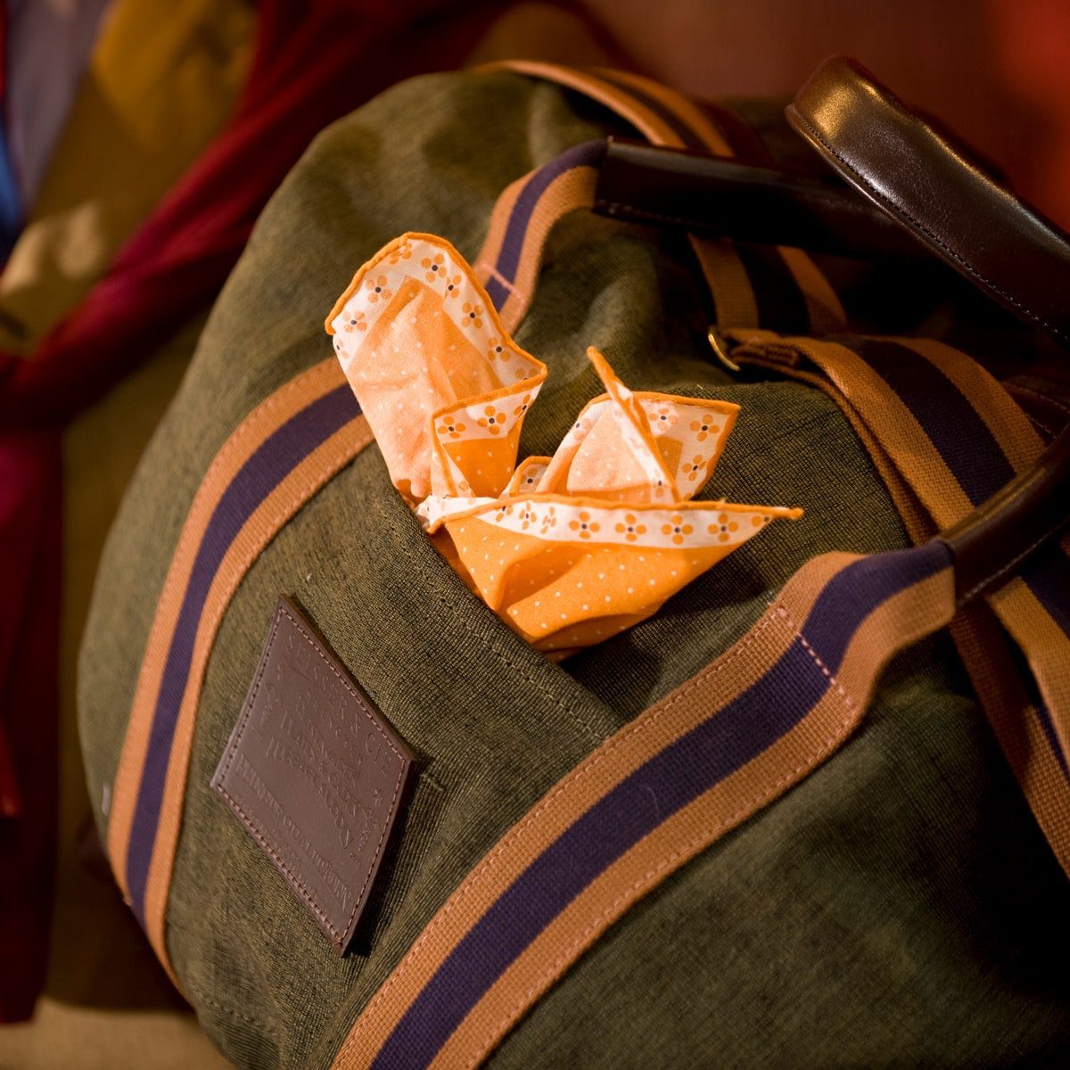 Nahaufnahme einer grün und braun gestreiften Reisetasche mit zwei orange gemusterten Origami-Booten in der Vordertasche. Das Licht schafft eine warme, gemütliche Atmosphäre.