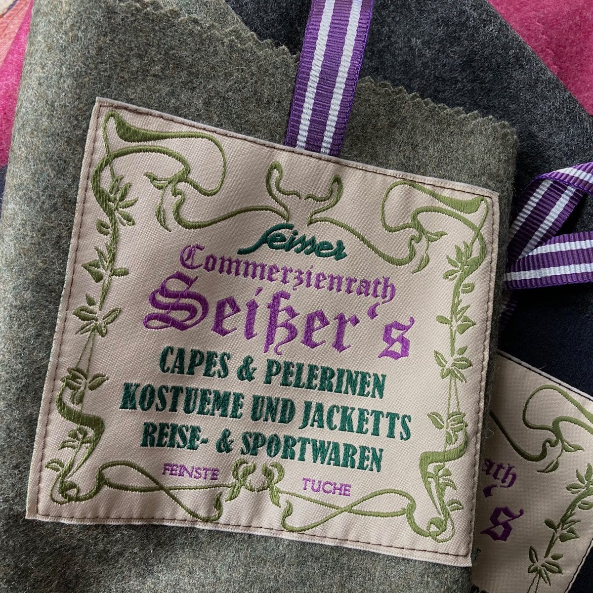Ein Stoffetikett mit violettem und grünem Text in deutscher Sprache, das für Umhänge, Jacken, Kostüme und Reisekleidung von "Seiker" wirbt, umrandet von einem verschnörkelten grünen Muster und mit einem violett gestreiften Band an einem Wollstoff befestigt.