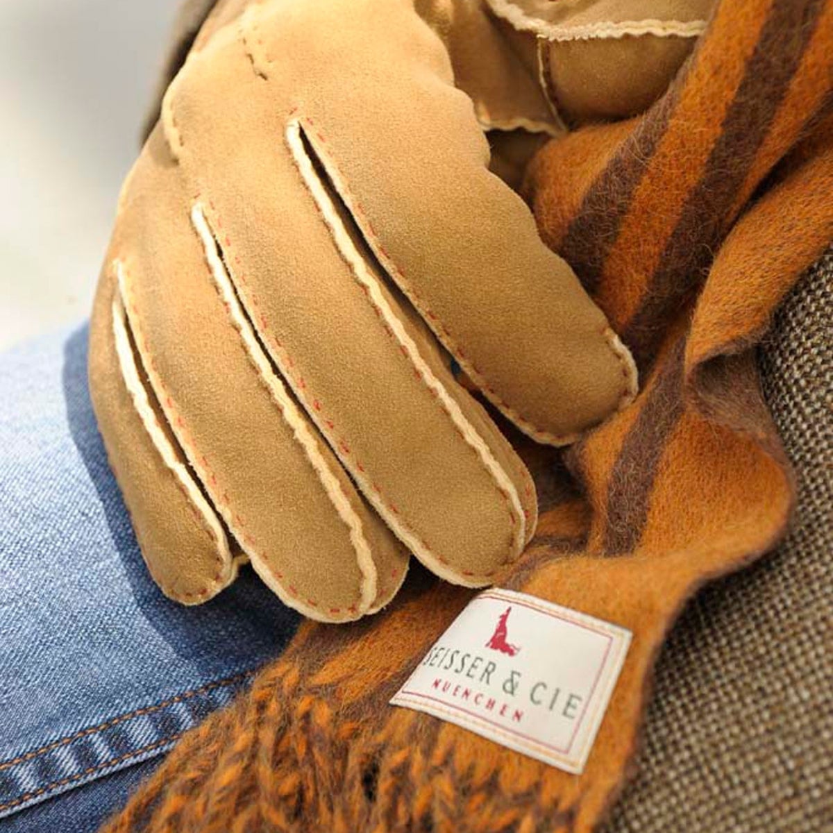 Nahaufnahme eines hellbraunen Wildlederhandschuhs, der auf einem Wollschal mit braunen und orangefarbenen Streifen ruht und ein Etikett mit der Aufschrift "HEISSER & CIE MÜNCHEN" zeigt. Ein Teil einer Tweedjacke und eine blaue Jeans sind ebenfalls zu sehen.