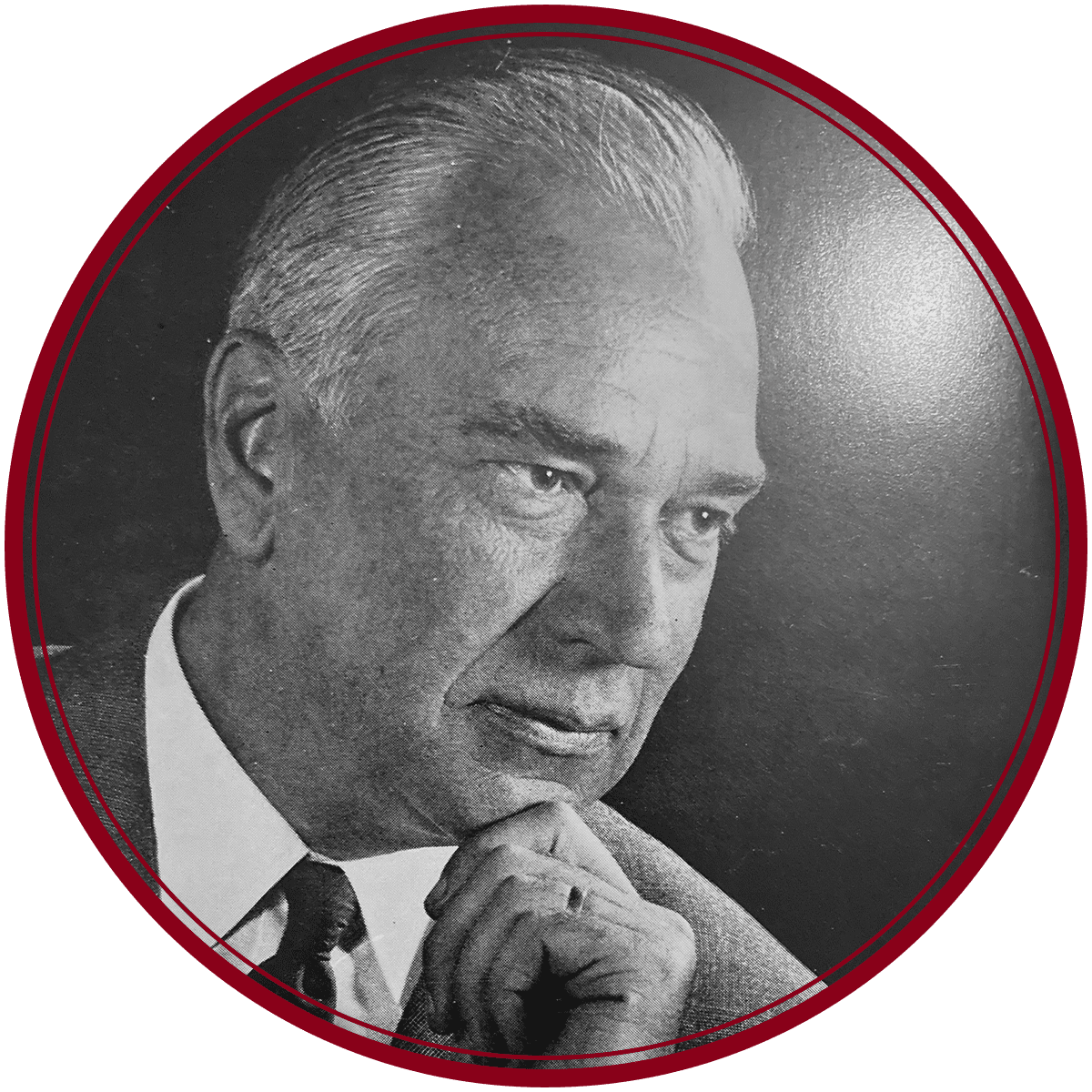 Schwarz-weißes Porträt eines älteren Mannes mit hellem Haar, Anzug und Krawatte, der sein Kinn in nachdenklicher Haltung auf die Hand stützt, umrahmt von einem roten kreisförmigen Rahmen.